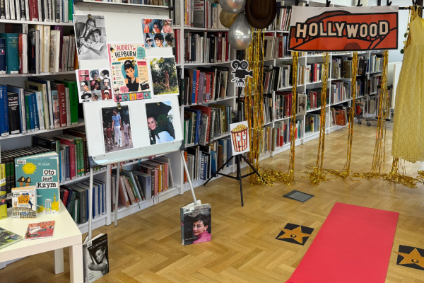 Zdjęcie dekoracji sali - czerwonego dywanu i gwiazd na podłodze, wystawki książek, tablicy ze zdjęciami Audrey Hepburn i złotej kurtyny z balonami i napisem Hollywood.