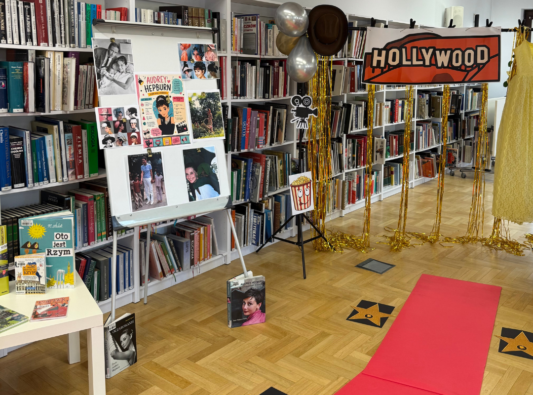 Zdjęcie dekoracji sali - czerwonego dywanu i gwiazd na podłodze, wystawki książek, tablicy ze zdjęciami Audrey Hepburn i złotej kurtyny z balonami i napisem Hollywood.