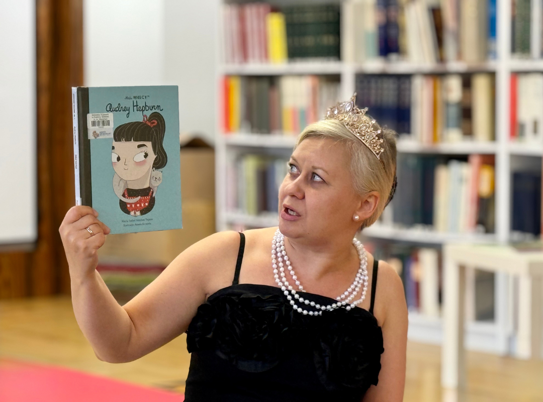 Zdjęcie prowadzącej warsztaty, ubranej w czarną sukienkę, perły i koronę, trzymającą książkę o Audrey Hepburn.