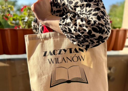 Osoba trzyma na ręku płócienną, kremową torbę z napisem "Zaczytany Wilanów" oraz infografiką w postaci otwartej książki.