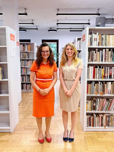 Zdjęcie Dyrektor biblioteki w pomarańczowej sukience (po lewej) oraz zastępcy Dyrektora w beżowej sukience (po prawej) na tle regałów.