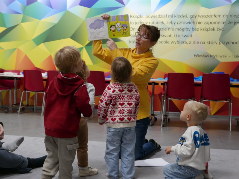 Zajęcia Klubu Małego Czytelnika. Bibliotekarka trzyma książkę i ją czyta. Wokół maluchy