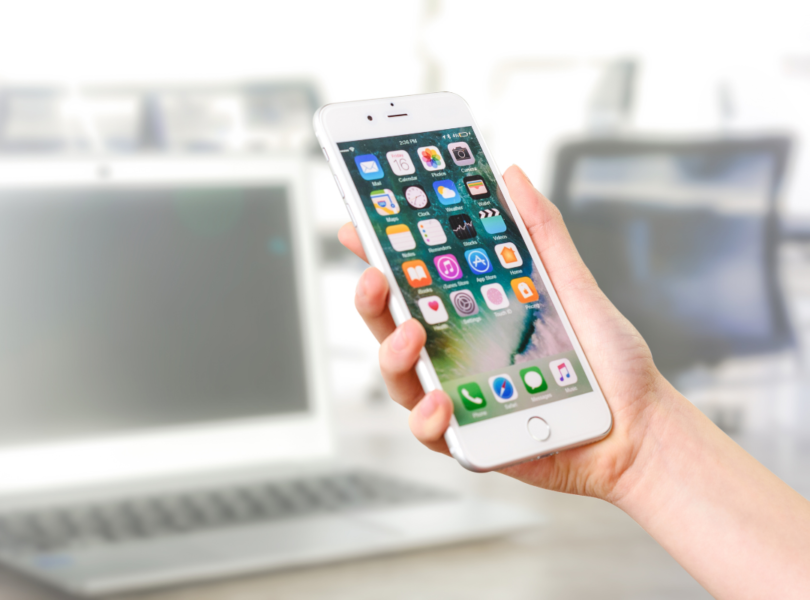 Na pierwszym planie ręka trzymająca smartfon, tło rozmazane, szare z zarysem laptopa i ekranu