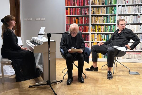 Pianistka siedzi wyprostowana przy pianie, w dwóch fotelach siedzą aktorzy, jeden czyta tekst