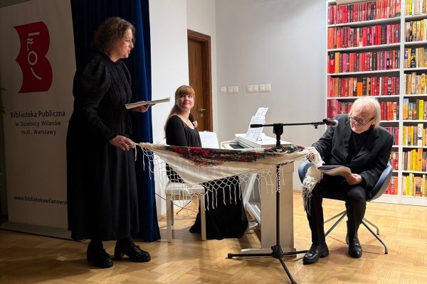 Pianistka siedzi przy białym pianinie, jeden z aktorów siedzi w fotelu i trzyma jeden róg chusty, drugi róg chusty trzyma stojąca obok aktorka